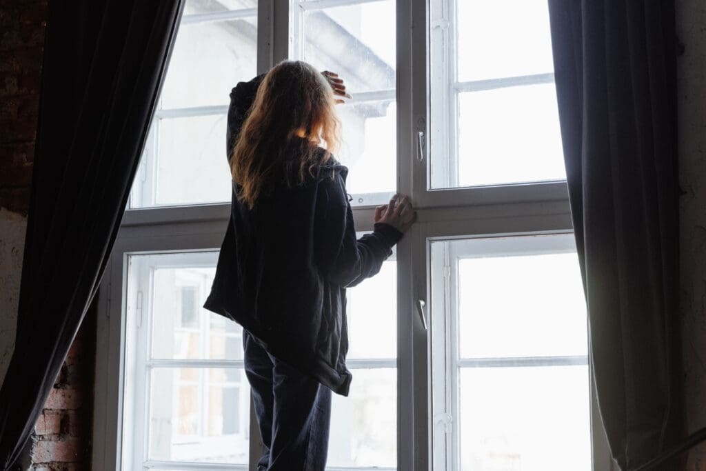 Back view of a woman standing at a window, looking outside with natural light streaming in
