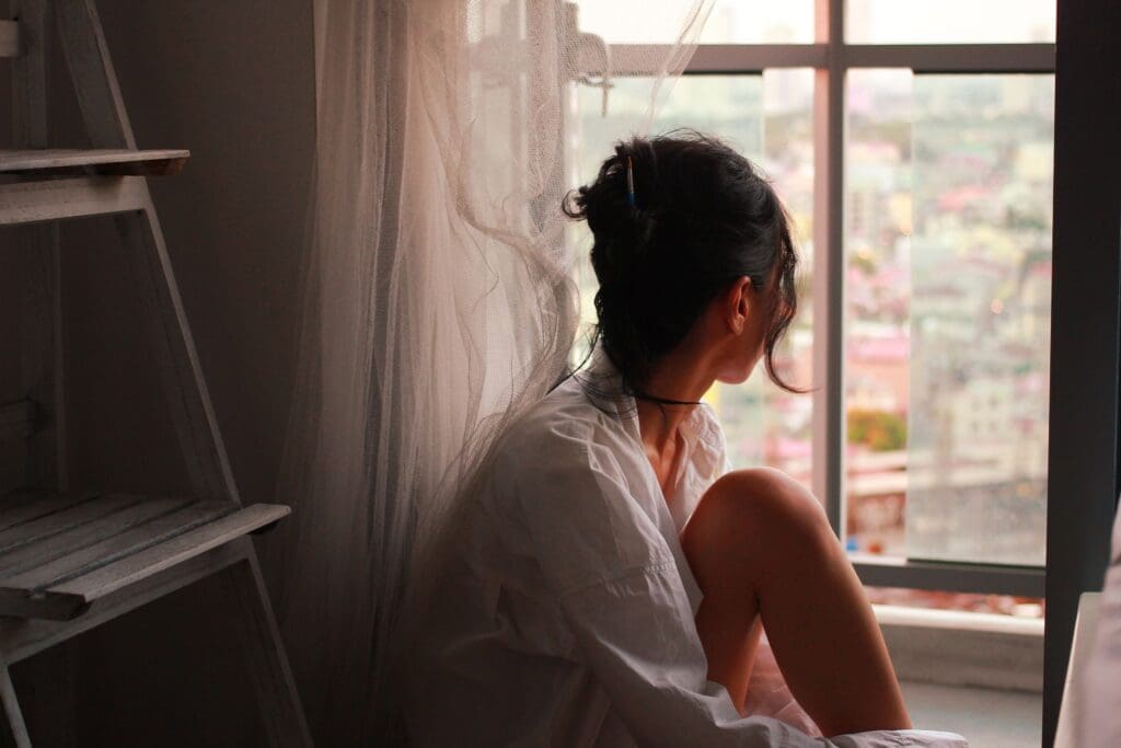 Woman sitting by a window in soft light, gazing outside with a distant, introspective expression