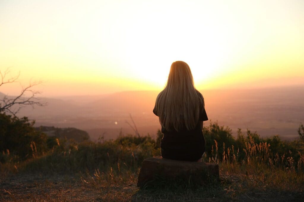Woman sitting alone on a hillside at sunset, viewed from behind, reflecting on her emotions