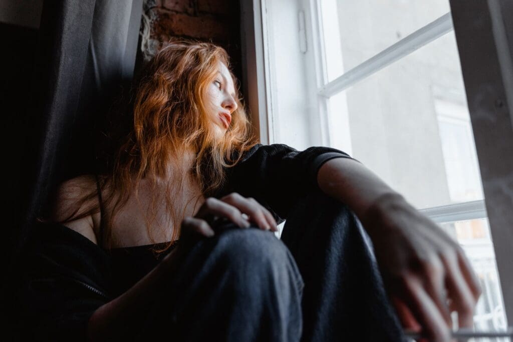Young woman with red hair sitting by a window, looking outside with a pensive and withdrawn expression