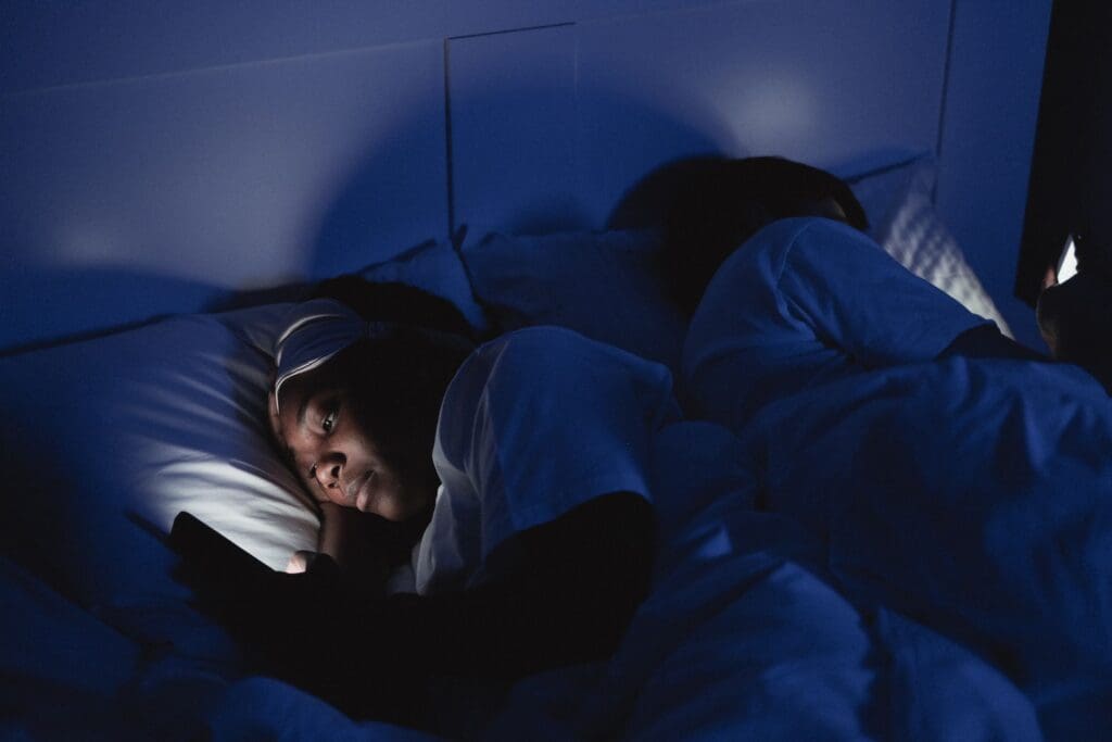 Person lying in bed at night scrolling on a phone in the dark while their partner sleeps beside them