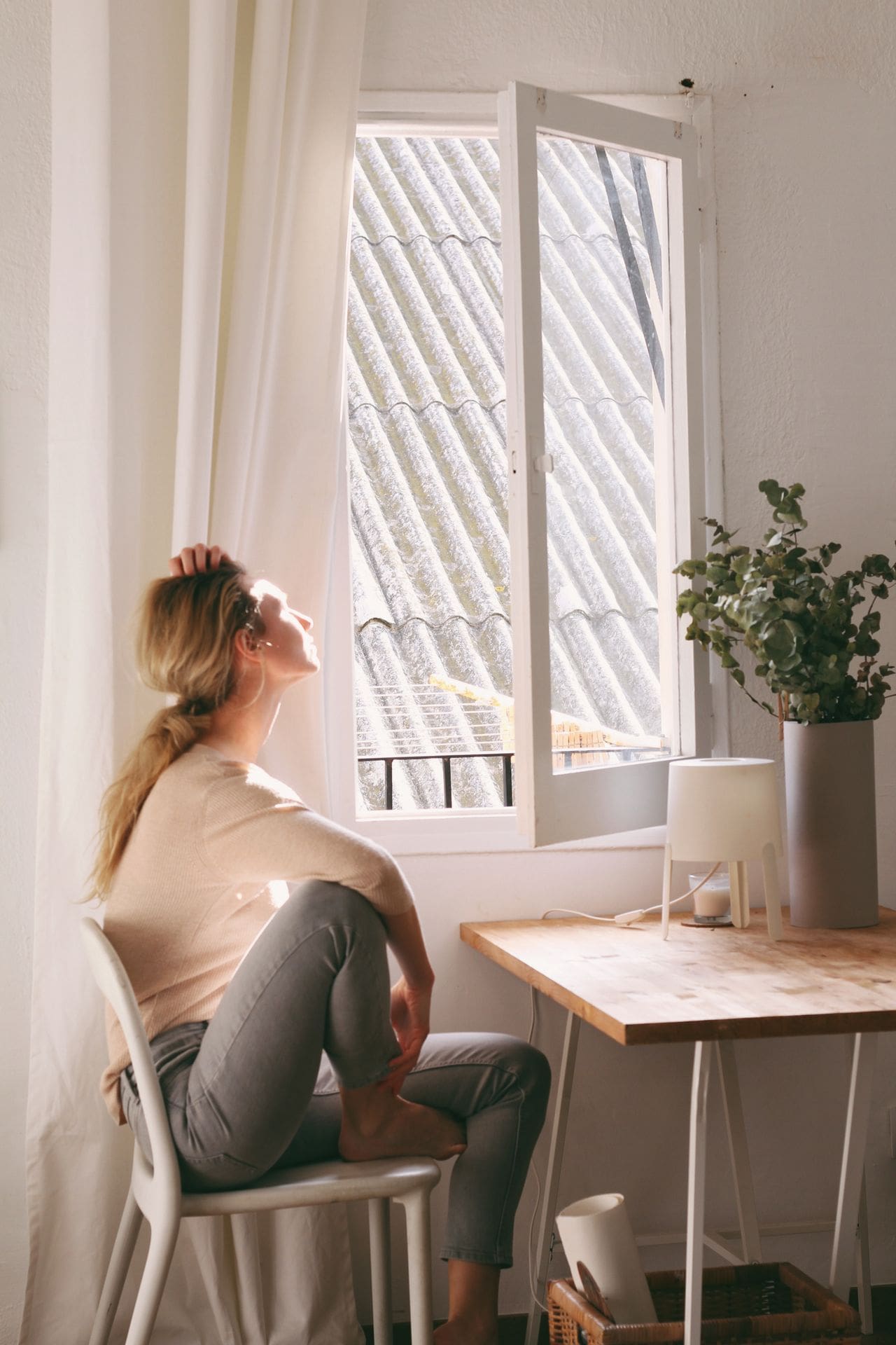 Woman sitting by a sunlit window in a calm, bright interior, taking a quiet moment to breathe and reflect