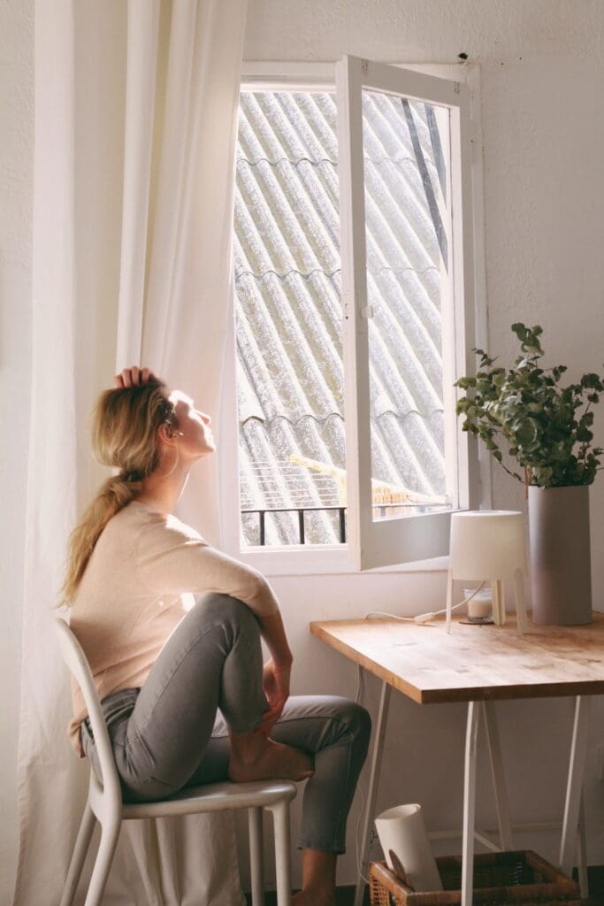 Woman sitting by an open window with eyes closed, face turned toward the sun, in a calm and quiet moment.