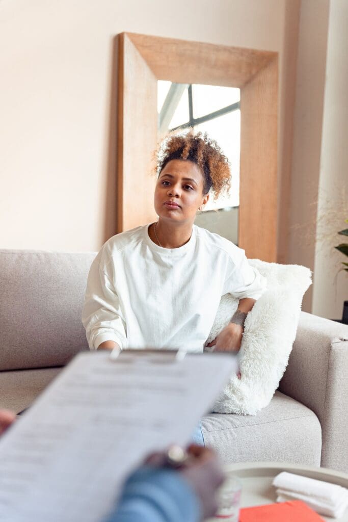 Woman sitting on a sofa during a therapy session, listening to a therapist holding a clipboard.