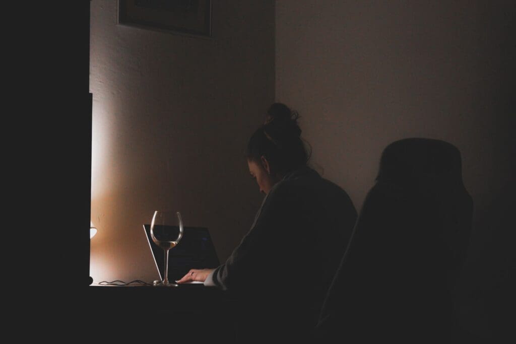 Woman working on a laptop late at night in a dark room, a glass of wine beside her.
