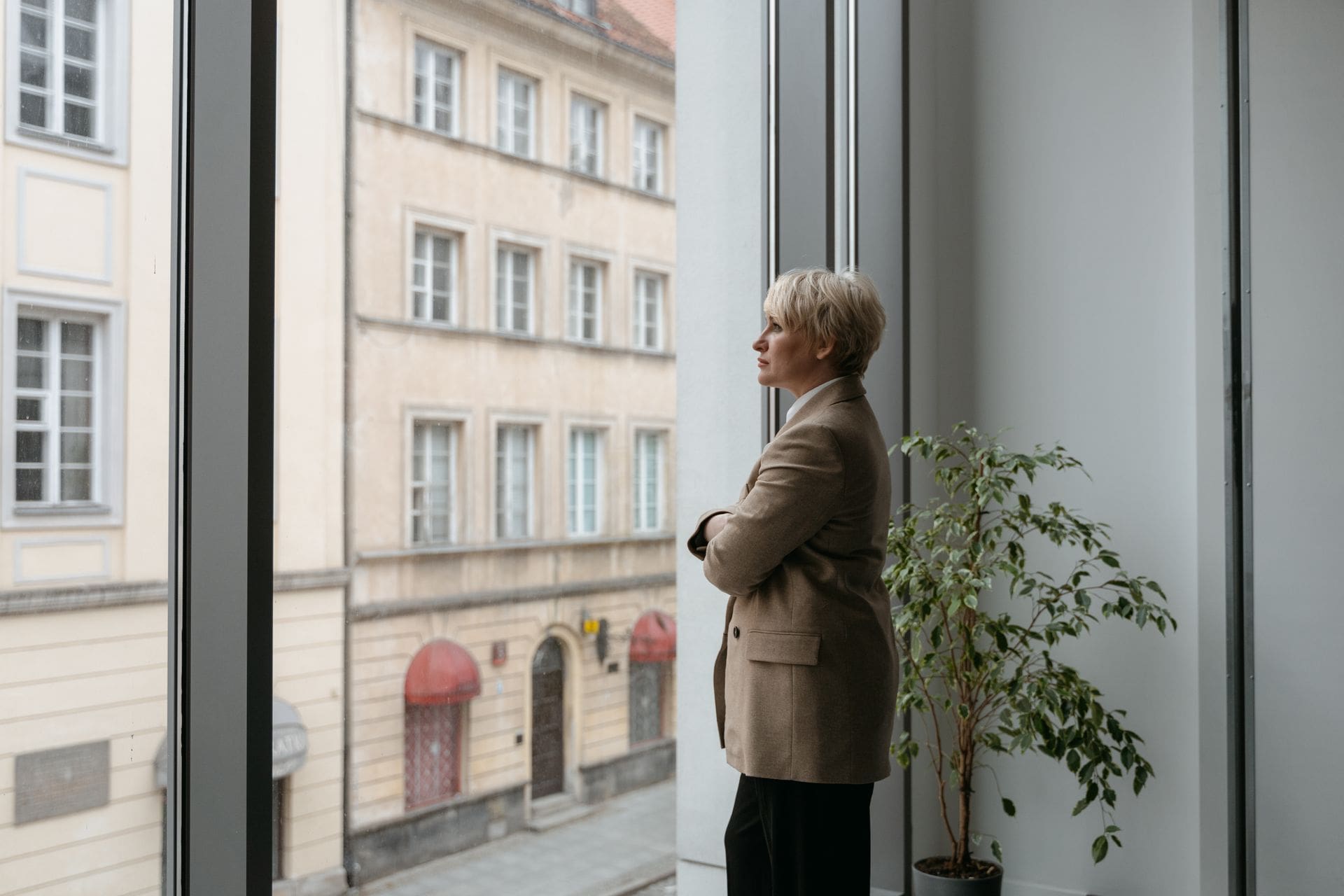 Woman in a blazer with arms crossed, looking out an office window, reflecting on process addiction and numbing behaviors.