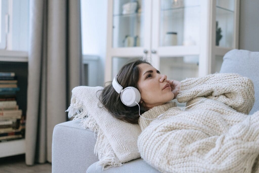 Woman lying on a sofa in a cozy sweater, listening to music with headphones, relaxed and present.