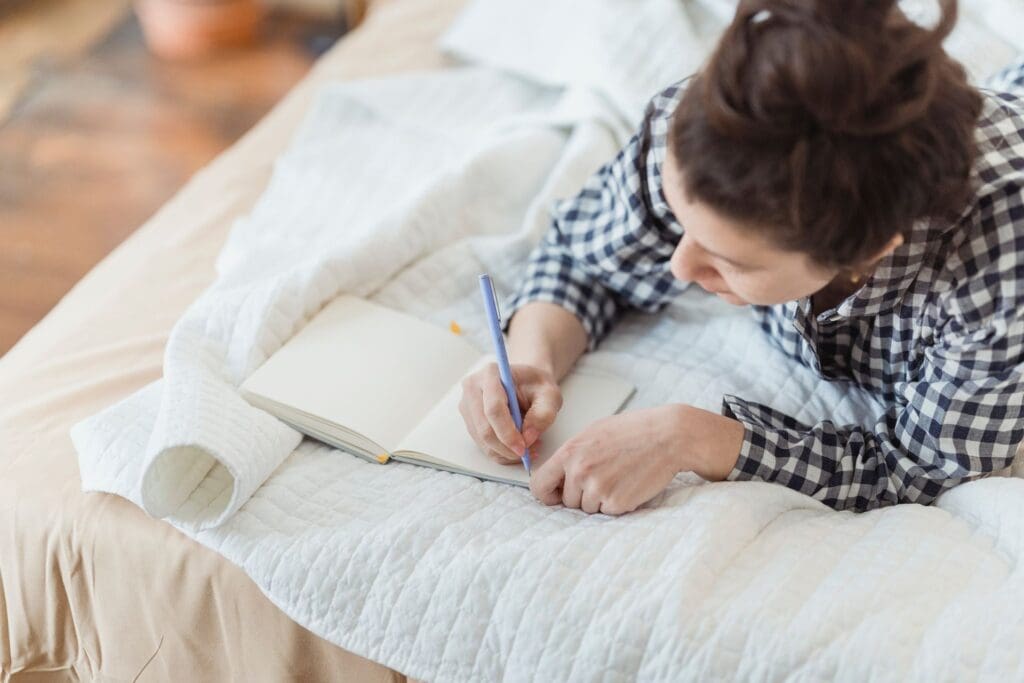 Woman lying on a bed writing in an open journal, taking quiet time for reflection.