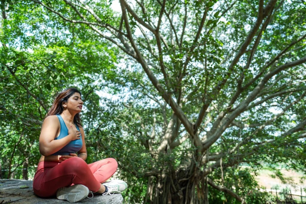 Woman sitting cross-legged under a large tree, eyes closed, one hand on her chest and one on her belly, practicing deep breathing.